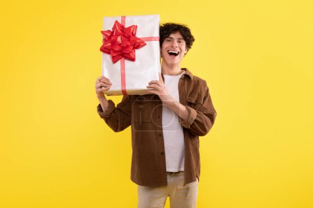 Téléchargez les photos : Moments de vacances. Joyful Guy posant avec une boîte-cadeau célébrant les fêtes et les anniversaires debout sur fond jaune studio. Offre de livraison publicitaire Jeune homme tenant présent, souriant à la caméra - en image libre de droit