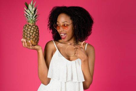 Téléchargez les photos : Satisfait millénaire afro-américain dame bouclée avec bouche ouverte et lunettes de soleil pointant du doigt à l'ananas isolé sur fond rose, studio. Plaisir d'été, détente et vacances - en image libre de droit