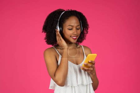 Foto de Feliz millennial afroamericana rizado dama en auriculares inalámbricos escuchando música, escribiendo en el teléfono inteligente aislado en el fondo rosa, estudio. Aplicación de audio, diversión, oferta y anuncio - Imagen libre de derechos