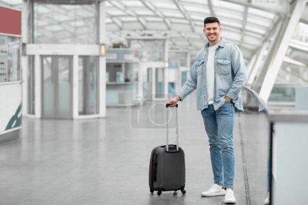 Téléchargez les photos : Offre de voyage pour Globetrotter. Homme voyageur posant avec valise bagages debout souriant à la caméra à l'aéroport intérieur. Prise de vue complète d'un jeune passager en attente de vol. Espace de copie - en image libre de droit
