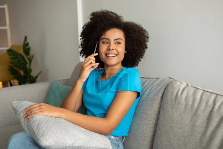 Photo for Laughing millennial curly black lady calling by smartphone,talking, enjoy gossip and good news, in cozy living room interior. Relax, device for communication remotely at home - Royalty Free Image