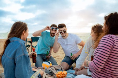 Foto de Felices amigos milenarios europeos y árabes en gafas de sol se divierten disfrutando del tiempo libre con botellas de cerveza en el picnic en la playa del océano con pelota y aperitivos. Vacaciones juntos, fiesta al aire libre - Imagen libre de derechos