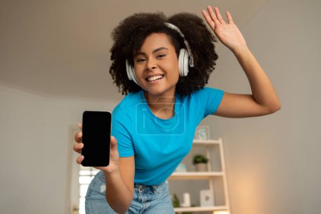 Photo for Positive young black curly woman in wireless headphones shows phone with blank screen, recommends audio app, enjoy free time alone, dancing and listen music in living room interior at home - Royalty Free Image