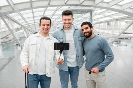 Foto de Exploradores emocionados. Tres viajeros hombres haciendo selfie en el teléfono móvil compartir momentos de viaje feliz en línea, posando abrazo en el aeropuerto moderno interior. Tech Fun And Tourism Concept - Imagen libre de derechos