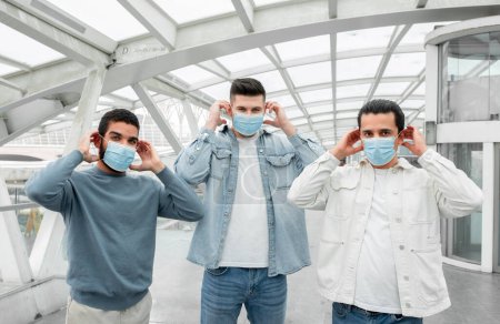 Téléchargez les photos : Trois touristes les gars portant des masques protecteurs médicaux pour la prévention du virus debout posant dans l'aéroport moderne à l'intérieur, regardant la caméra. Concept Voyage et soins de santé - en image libre de droit