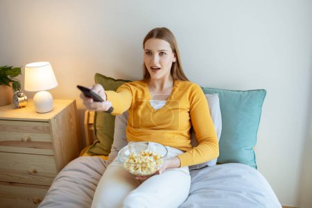 Photo for TV Entertainment. Cheerful Young Lady Watching Television Enjoying Favorite Shows And Movies, Eating Popcorn Sitting In Cozy Bedroom Indoor. Excited Woman Switching Channels With Remote Controller - Royalty Free Image