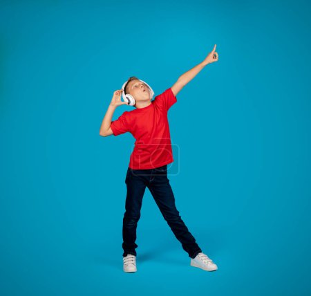Cheerful Caucasian Boy Wearing Wireless Headphones Listening Music And Dancing, Preteen Male Child Having Fun While Standing Over Blue Background In Studio, Full Length Shot, Copy Space