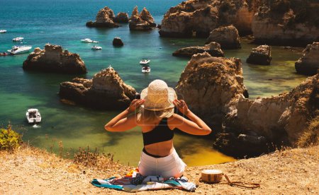 Téléchargez les photos : Vacances au Portugal. Vue arrière d'une touriste assise sur une falaise portant un chapeau d'été, regardant la vue sur l'océan, des rochers et des bateaux dans l'eau. Dame méconnaissable jouissant d'une journée ensoleillée au bord de la mer à l'extérieur - en image libre de droit