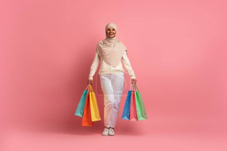 Téléchargez les photos : Pleine longueur prise de vue de femme musulmane heureuse marchant avec des sacs à provisions lumineux sur fond rose studio, sourire arabe Shopaholic Lady In Hijab portant des achats, profiter des ventes saisonnières, Copier l'espace - en image libre de droit