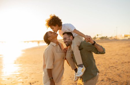 Photo for Beachside bliss. Family of three spending free time by sea, walking on the beach and enjoying family bonding time outdoors, free space. Father carrying son on shoulders - Royalty Free Image