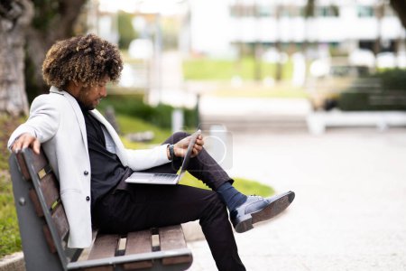 Photo for Black businessman in suit using laptop while sitting on bench near modern office building, side view, free space. Conference due social distance, new normal concept - Royalty Free Image