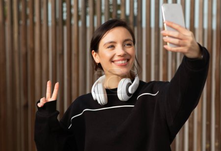 Foto de Feliz millennial mujer atleta europea en ropa deportiva tomar selfie en el teléfono, señal de paz con la mano en la ciudad, al aire libre. Entrenamiento de fitness y pilates, blog de cuidado corporal, aplicación de pérdida de peso y estilo de vida activo - Imagen libre de derechos