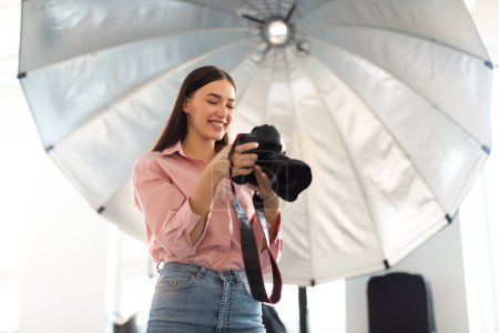 Young photographer lady working with professional camera in front of reflective umbrella, taking photos and enjoying her work in modern photostudio, copy space