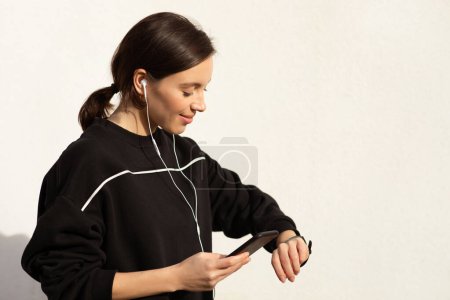 Photo for Positive millennial european woman athlete in sportswear and headphones enjoy music and workout, checks heart rate on phone and fitness tracker on white wall background, outdoor. Body care, fitness - Royalty Free Image