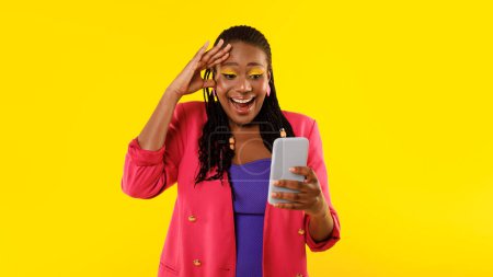 Téléchargez les photos : Femme afro-américaine excitée regardant Smartphone dans l'excitation, en utilisant des applications debout sur fond jaune Studio. Jeune femme faisant défiler les médias sociaux Découvrir le Web par téléphone cellulaire. Panorama - en image libre de droit