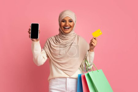Photo for Shopping App. Happy Muslim Lady Holding Smartphone, Shopper Bags And Credit Card, Smiling Islamic Female In Hijab Showing Mobile Phone With Blank Empty Screen, Recommending Online Store, Mockup - Royalty Free Image