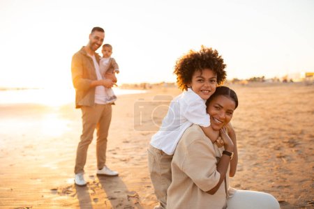 Photo for Beachside bonds. Mother and son embracing and smiling at camera, father with little son standing on background. Parents spending time with sons outdoors - Royalty Free Image