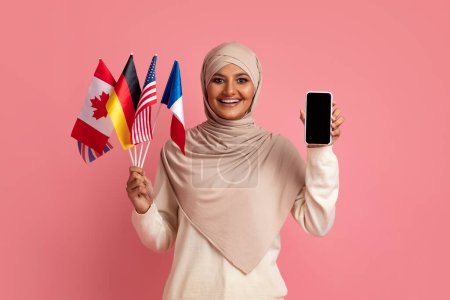 Téléchargez les photos : Heureuse femme musulmane en hijab tenant divers drapeaux et smartphone vierge avec écran noir, souriante dame islamique apprenant des langues étrangères en ligne, en utilisant l'application mobile, fond rose, maquette - en image libre de droit