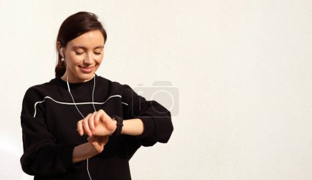 Téléchargez les photos : La jeune femme caucasienne positive avec écouteurs aime la musique, se repose de l'entraînement, vérifie le pouls sur le tracker sur fond de mur blanc. Sport, fitness et audio app, perte de poids, soins de santé en ville - en image libre de droit
