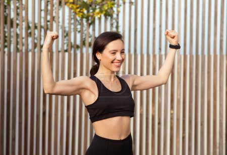 Positive millennial european woman athlete champion show muscles biceps on hands, celebrating victory in competition in city, outdoor. Fitness and strength training, body care
