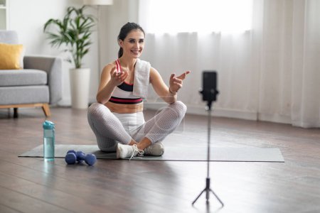 Photo for Well-fit sporty young brunette woman wearing sportswear sitting on yoga mat, looking at cell phone camera, talking and gesturing, fitness coach recording online lesson, home interior, copy space - Royalty Free Image