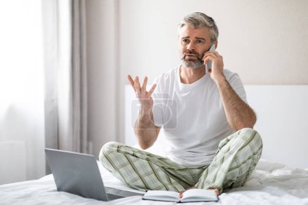 Téléchargez les photos : Irrité en colère beau aux cheveux gris homme d'âge moyen portant un pyjama assis sur le lit à la maison, à l'aide d'un ordinateur portable, avoir une conversation téléphonique et des gestes, avoir des disputes, copier l'espace - en image libre de droit