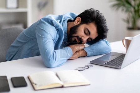 Téléchargez les photos : Concept surmenage. Fatigué jeune homme indien sieste au bureau avec ordinateur portable, Est Homme freelancer épuisé après avoir travaillé sur l'ordinateur au bureau à domicile, Placer la tête sur les mains, Gros plan - en image libre de droit