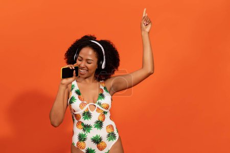 Téléchargez les photos : Émotionnelle heureuse millénaire attrayante dame afro-américaine en maillot de bain à la mode s'amuser pendant les vacances d'été, à l'aide d'écouteurs, téléphone portable, chant, danse sur fond orange, espace de copie - en image libre de droit