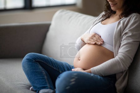 Foto de Hora prenatal. Mujer embarazada irreconocible sentada en el sofá y abrazando su vientre, sonriente joven embarazada que se relaja en casa, cariñosa barriga, primer plano con espacio libre - Imagen libre de derechos