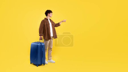 Téléchargez les photos : Offre de voyage Publicité. Jeune homme avec sac à valise bleue pointant du doigt à côté de l'espace de copie, service de réservation de billets publicitaires debout en studio sur fond jaune. Panorama - en image libre de droit