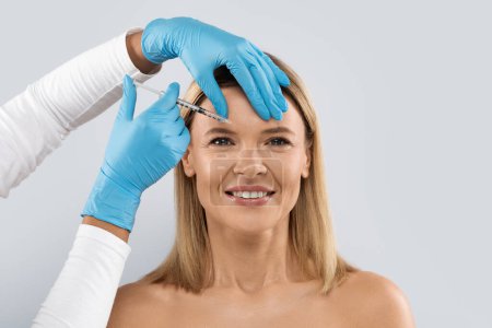 Photo for Beautiful half-naked blonde middle aged woman gets cosmetic injection between eyebrows. Beautician hands making hyaluronic acid injection for glabella facial rejuvenation procedure, studio background - Royalty Free Image