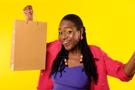 Foto de Shopaholics Joy. Alegre dama afroamericana celebrando compras exitosas mostrando bolsa de papel sobre fondo amarillo. Studio Shot. Oferta de ventas Concepto de publicidad - Imagen libre de derechos