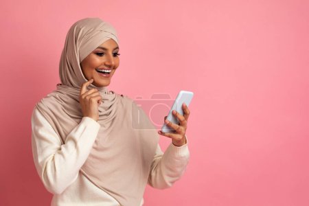 Téléchargez les photos : Souriant jeune femme musulmane regardant l'écran du smartphone avec intérêt, heureuse dame islamique tenant téléphone portable et toucher le menton, bénéficiant d'une application moderne ou d'une offre en ligne tout en se tenant debout sur fond rose - en image libre de droit