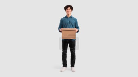 Photo for Delivery Offer. Happy Guy Holding Cardboard Moving Box Smiling To Camera Standing In Studio Over White Background. Man Carrying Parcel Package Advertising Shipment Service. Panorama - Royalty Free Image