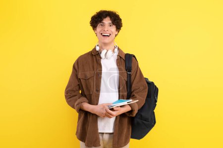 Photo for Joyful Learner. Excited Student Guy With Headphones And Textbooks Expressing Positivity Advertising University Education, Posing Standing With Backpack On Yellow Background In Studio - Royalty Free Image