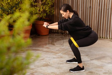 Téléchargez les photos : Heureuse athlète européenne millénaire en tenue de sport squat avec élastique ajusté en ville, en plein air. Fitness et pilates entraînement, équipement de soins du corps, perte de poids et mode de vie actif - en image libre de droit