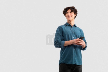 Photo for Digital Communication. Cheerful Guy Browsing Social Media On His Smartphone, Texting And Websurfing Standing On White Studio Background, Looking Aside At Empty Space For Text - Royalty Free Image