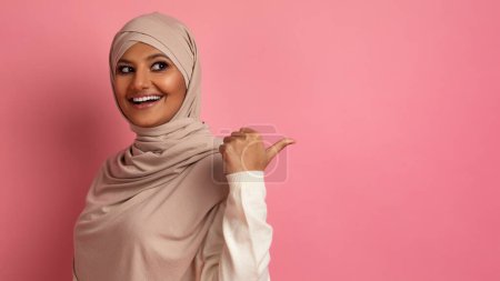 Téléchargez les photos : Belle Promo. Femme musulmane heureuse dans le hijab pointant vers l'espace de copie derrière son dos avec le pouce levé et souriant, dame arabe excitée montrant la place libre pour l'annonce, debout sur fond de studio rose, Panorama - en image libre de droit