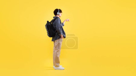 Photo for Side View Of Young Man With Backpack Wearing Headphones Listening To Music And Pressing Invisible Button Or Showing Free Space Standing On Yellow Studio Background. Panorama - Royalty Free Image