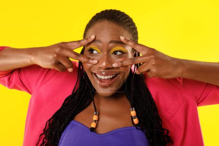 Photo for Victory And Peace Concept. Portrait Of Cheerful Black Lady Gesturing V Signs Near Eyes Posing With Bright Makeup Having Fun Over Yellow Background, Looking Aside. Studio Shot - Royalty Free Image