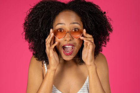 Foto de Sonriente mujer rizada afroamericana milenaria sorprendida con boca abierta y gafas de sol aisladas sobre fondo rosa, estudio. Vacaciones, diversión de verano, relax y vacaciones, moda - Imagen libre de derechos