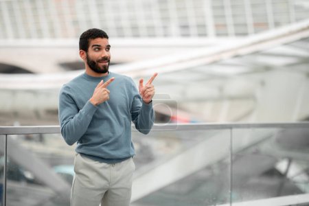Photo for Travel Tickets Offer. Excited Arabic Guy Pointing Fingers Aside At Free Space Advertising Transportation Service Standing At Modern Airport Departure Terminal Indoor. Look There Concept - Royalty Free Image