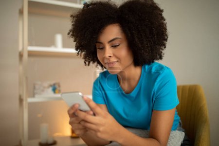 Photo for Glad millennial curly black lady typing on smartphone, watching video, surfing in internet in living room interior. Device and social networks, blog and chat, communication remotely at home - Royalty Free Image