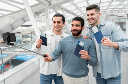 Foto de Momento de aventura de viaje. Tres turistas alegres hombres haciendo selfie por teléfono mostrando sus boletos de embarque posando en el aeropuerto moderno y divirtiéndose juntos. Concepto de blogueo de vacaciones - Imagen libre de derechos