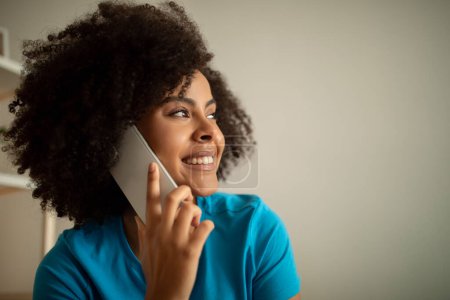 Photo for Smiling young black curly woman calling by smartphone, talking to friend, enjoy free time, look at copy space in living room interior, close up. Sale, good news, communication, gossip at home - Royalty Free Image