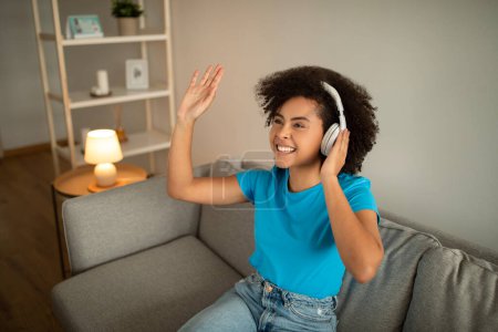 Satisfied millennial curly black lady in wireless headphones waving hand, enjoys free time on sofa in cozy living room interior. Housewife listen music, audio app, video call