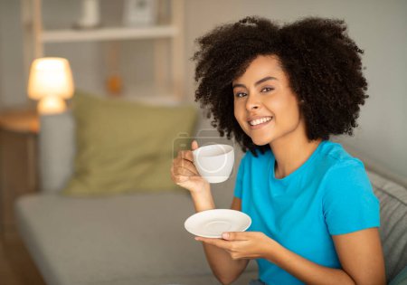 Photo for Good morning and fresh drink. Cheerful young black curly woman drinks cup of hot coffee on sofa, enjoy free time alone in evening, relax in living room interior. Tea and rest at home, ad and offer - Royalty Free Image