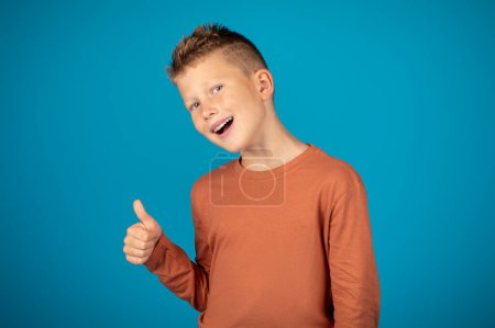 Photo for I Like It. Cheerful Caucasian Boy Showing Thumb Up At Camera, Positive Tween Male Child Recommending Something, Gesturing Sign Of Approval While Standing Over Blue Studio Background, Copy Space - Royalty Free Image