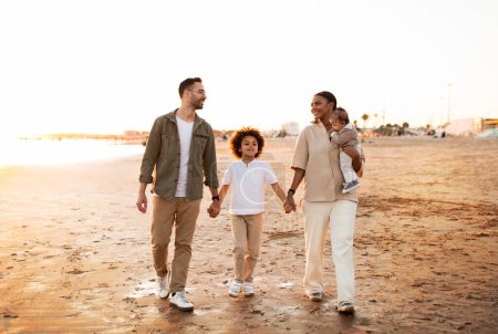 Photo for Love on the shoreline. Family walking on the beach with two sons, holding hands and smiling, enjoying evening by seaside. Diverse parents spending time with children together outdoors - Royalty Free Image