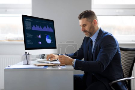 Téléchargez les photos : Homme d'affaires d'âge moyen travaillant sur ordinateur, assis devant moniteur avec des statistiques de trading en ligne sur le lieu de travail au bureau. Homme entrepreneur écrit dans un carnet - en image libre de droit